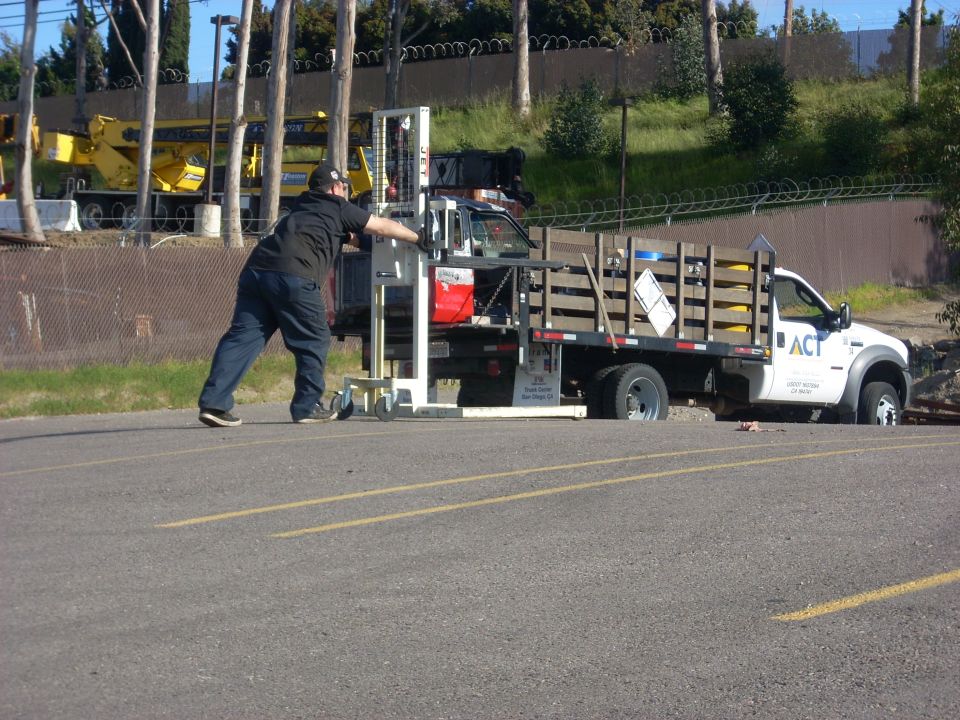 Man pushes a forklift onto the bed of a white flatbed truck with 'ACT' logo. Another truck is visible in the background