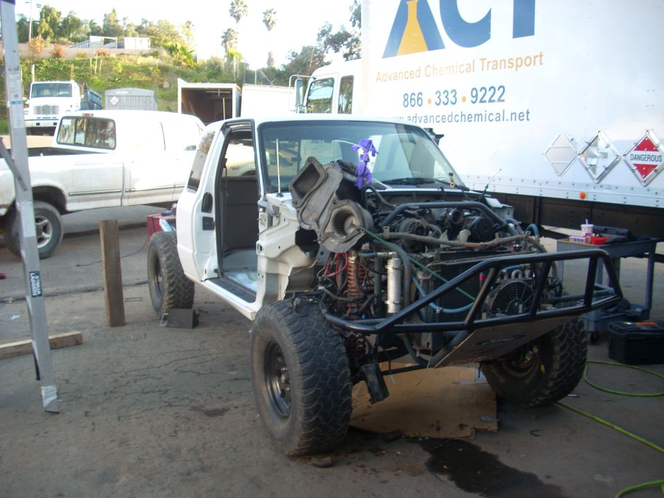White pickup truck with front end removed, showing engine and chassis. Another white pickup truck is parked behind it