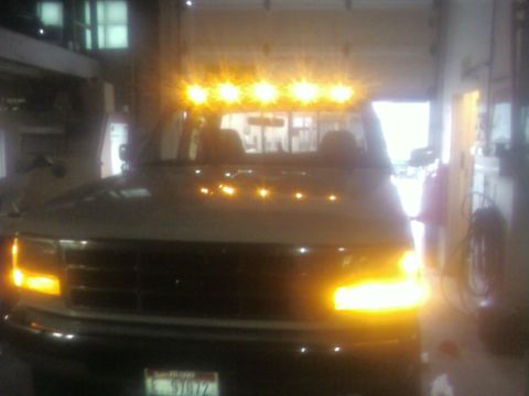 1989 Ford Bronco - Front view of a light-colored Ford SUV parked indoors in a garage, with a roof-mounted amber cab light bar illuminated and both headlights/…