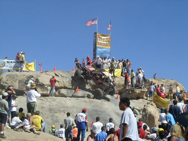 Off-road buggy climbing a steep, rocky incline with spectators watching. American flags fly on a tall sign in the background
