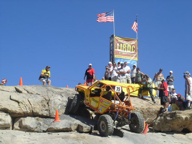Yellow off-road buggy with number 75 on a rocky incline, with spectators and flags