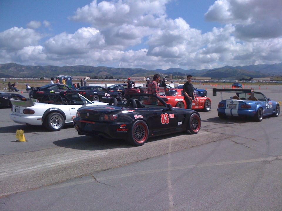 Black Honda S2000 #83 SSM autocross car staged beside white and blue Miatas on an airfield lot