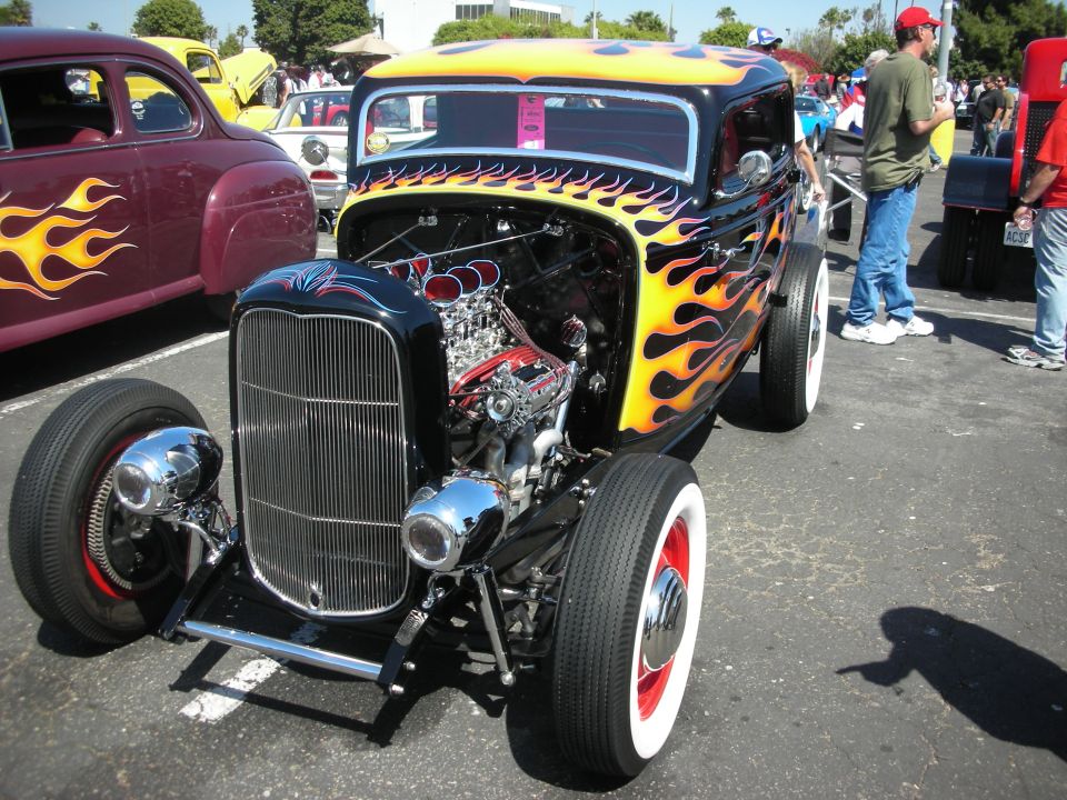 Hot rod with flames painted on it, exposed engine, and whitewall tires. Another car with flames is parked next to it