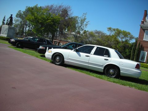 2004 Ford Crown Victoria - Three police cars are parked in a row on a sunny day with a brick building in the background.