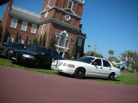 2004 Ford Crown Victoria - Two black and one white Ford Crown Victoria police cars are parked in front of a brick building.