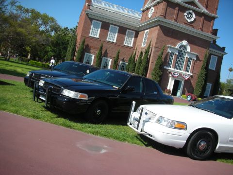2004 Ford Crown Victoria - Three Ford Crown Victoria police cars are parked on the lawn in front of a brick building with a white-trimmed facade.