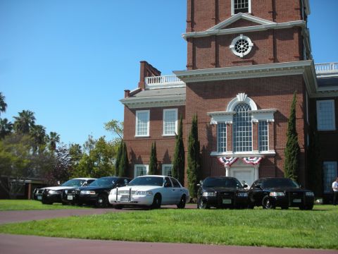 2004 Ford Crown Victoria - A line of police cruisers are parked on the grass in front of a large brick building with a clock tower.