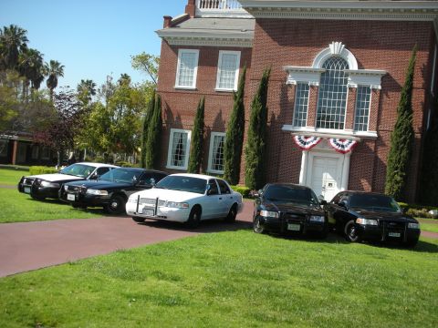 2004 Ford Crown Victoria - Several police sedans are lined up on a lawn in front of a large brick building with white trim.