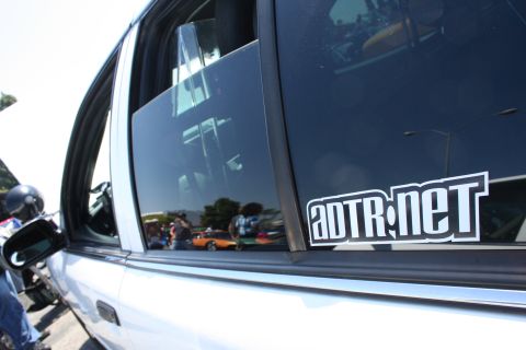 2004 Ford Crown Victoria - Car door with 'adtr.net' sticker, reflections show other vehicles and people at an event.