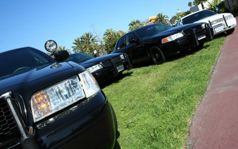 2004 Ford Crown Victoria - Multiple police cruisers are lined up on a grassy area under a clear blue sky.