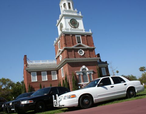 2004 Ford Crown Victoria - Two Ford Crown Victoria police cars are parked on a grassy area in front of a large brick building with a clock tower under a clear blue sk…