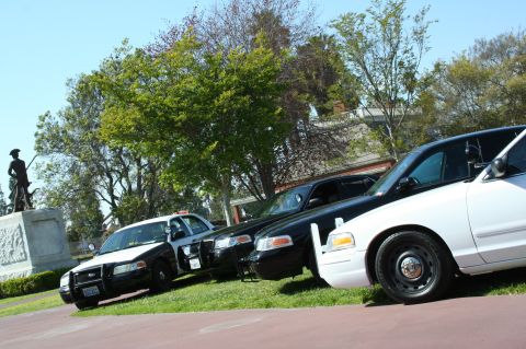 2004 Ford Crown Victoria - Several police vehicles, including a white and black Crown Victoria, are parked on a grassy area under a clear blue sky.
