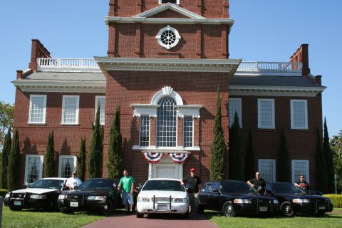 2004 Ford Crown Victoria - Several police cruisers are lined up in front of a historic brick building on a sunny day.