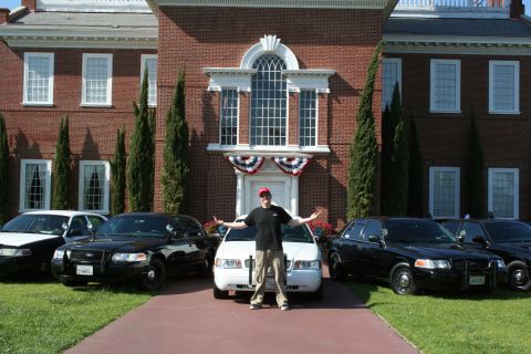 2004 Ford Crown Victoria - A man poses with multiple Ford Crown Victoria police cars in front of a large brick building.
