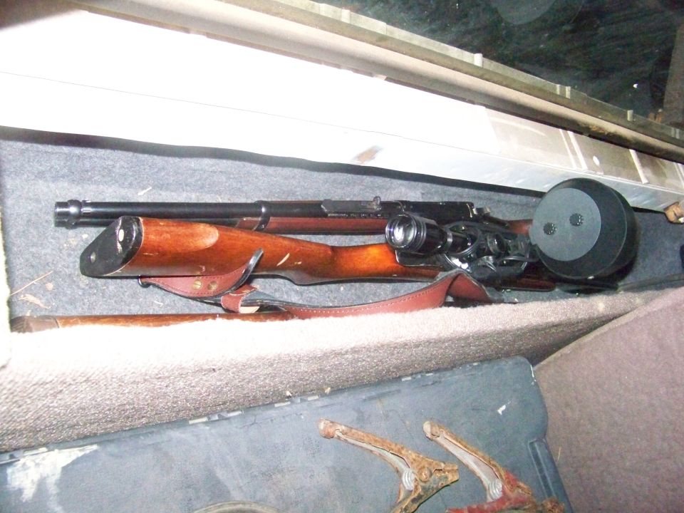 Rifle with wooden stock, scope, and drum magazine resting on car seat