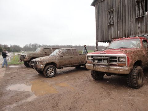 1979 Ford Bronco - Three pickup trucks are covered in mud, parked near a rustic wooden barn on an overcast day.