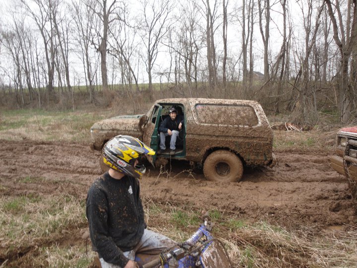 Muddy SUV stuck in a field with a dirt bike in the foreground. Two people are in the SUV