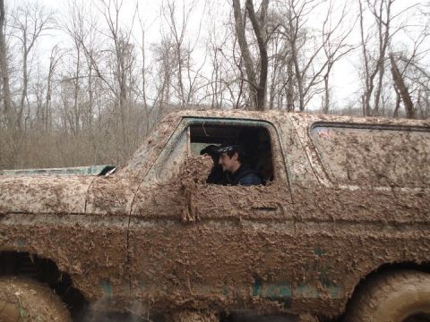 1979 Ford Bronco - A 1979 Ford Bronco is heavily caked in mud, with a driver visible through the window, navigating an off-road trail.