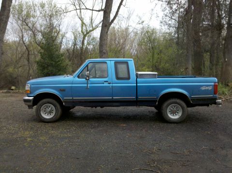 1992 Ford F-150 - A blue pickup truck is parked on a gravel surface with trees in the background.