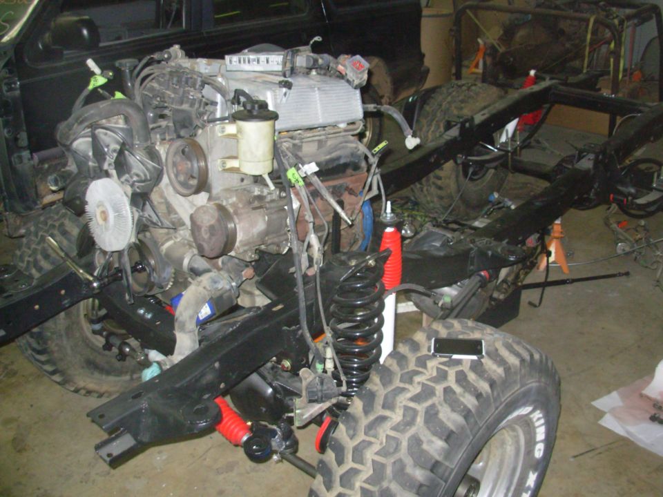 Truck frame and front suspension with engine installed, red coil-over shock and mud tires during a build