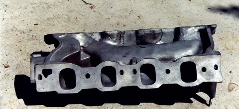 1973 Ford Mustang - An exhaust manifold sits on a concrete surface, showing its intake ports and some casting marks.