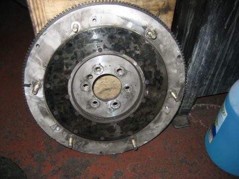 1998 Ford Crown Victoria - used engine flywheel with its toothed ring gear and bolted center hub, propped against wood on a shop floor.