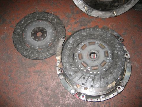 1998 Ford Crown Victoria - worn clutch disc beside a pressure plate with sprung center hub, laid on a concrete floor near a flywheel.