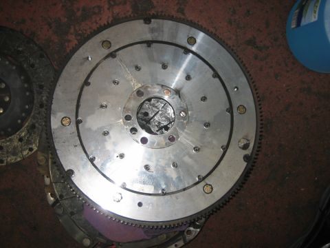 1998 Ford Crown Victoria - aluminum flywheel with a pressed-on steel starter ring gear and a bolted central crank flange sits on a red shop floor.