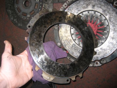 1998 Ford Crown Victoria - hand holds a worn metal clutch friction ring over a pile of clutch parts including pressure plates and discs on a shop floor.