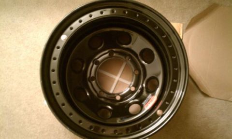 1990 Ford Bronco - Overhead view of a black steel wheel lying face-up on carpet next to an opened cardboard box, showing a five-lug bolt circle, six large rou…