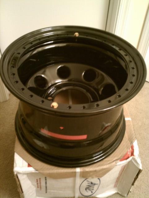 1990 Ford Bronco - A gloss black steel wheel with a beadlock-style outer ring drilled for a full circle of clamp bolts, sitting face-up on a cardboard shippin…