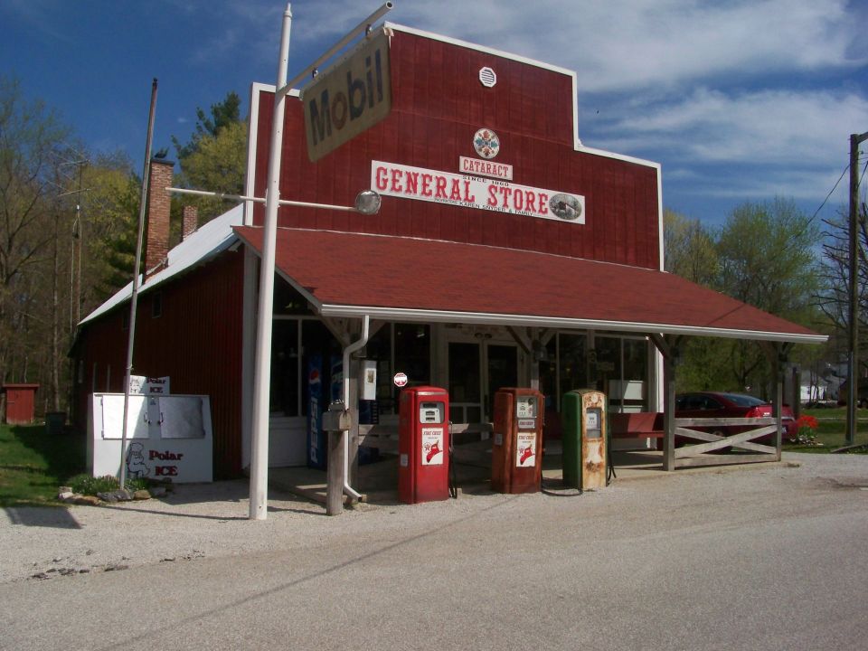 Red general store with a white roof and porch, three vintage gas pumps, and a red car parked under the porch