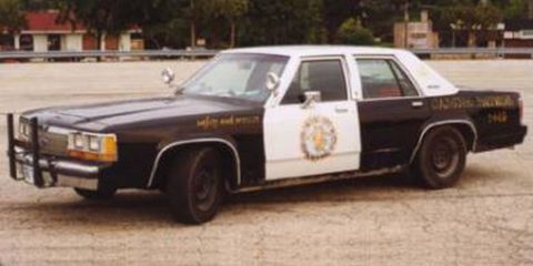 1991 Ford LTD Crown Victoria - Black-and-white Ford LTD Crown Victoria highway patrol sedan shot from the front three-quarter on a concrete lot, wearing a gold star door…