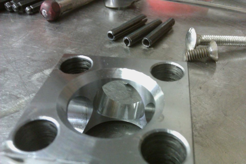 Machined metal part with four threaded holes, three pins, and two screws on a textured surface