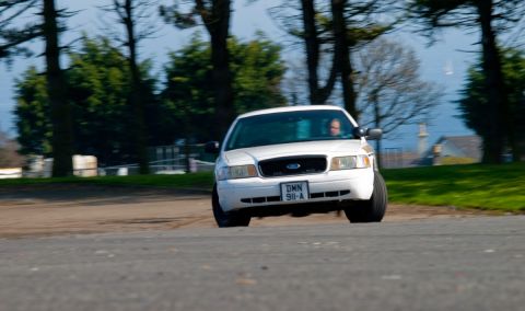 2001 Ford Crown Victoria - A white 2001 Ford Crown Victoria is captured in motion on a paved road, with trees and a distant building in the background.