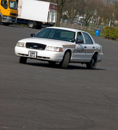 2001 Ford Crown Victoria - A white 2001 Ford Crown Victoria police car is seen from the front-right, with a yellow truck in the background.