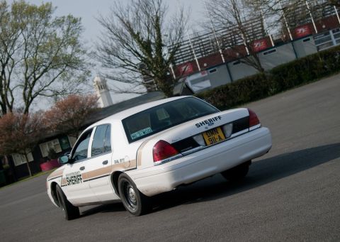 2001 Ford Crown Victoria - A sheriff's car is seen from the rear, driving on a paved surface with buildings and trees in the background.