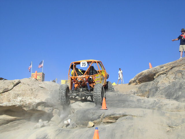 Orange off-road buggy with number 75 on roof climbing a rocky incline under a clear blue sky