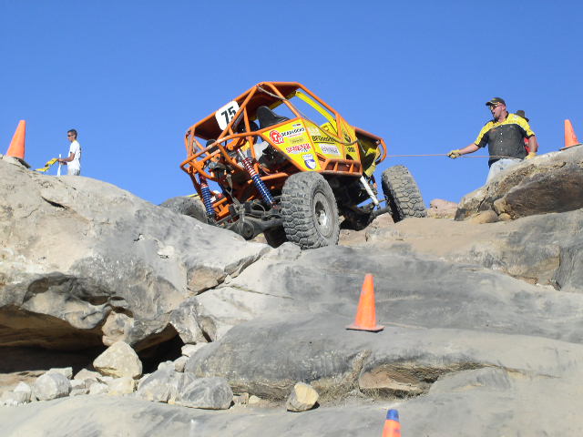 Off-road buggy with number 75 on it is being pulled up a rocky incline by a rope held by a man
