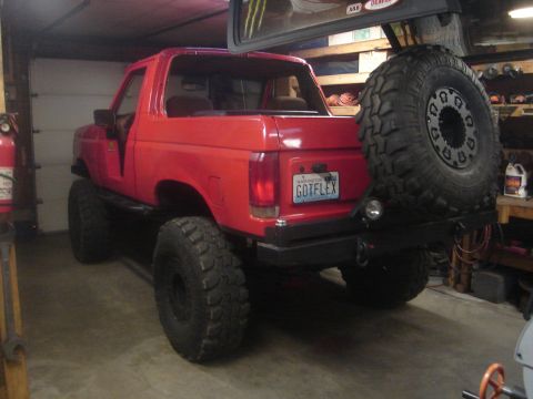 1989 Ford Bronco - Three-quarter rear view of a lifted red Ford Bronco in a garage with its doors and hardtop removed, the fiberglass top hoisted overhead. It…