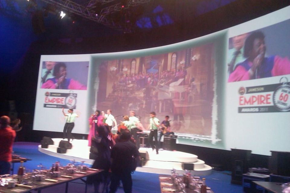 Stage performance with musicians and dancers in front of a large screen showing award ceremony footage