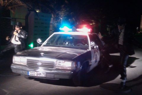 1991 Ford LTD Crown Victoria - A police car with its lights on is parked at night, with two men in dark suits and hats standing next to it.