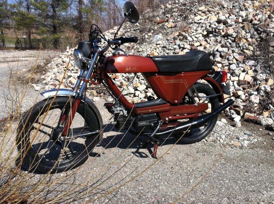 Red moped with black seat and chrome fender parked on gravel