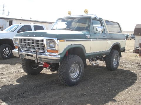 1979 Ford Bronco - A lifted Ford Bronco sits in a dirt lot, with a modern pickup truck nearby.