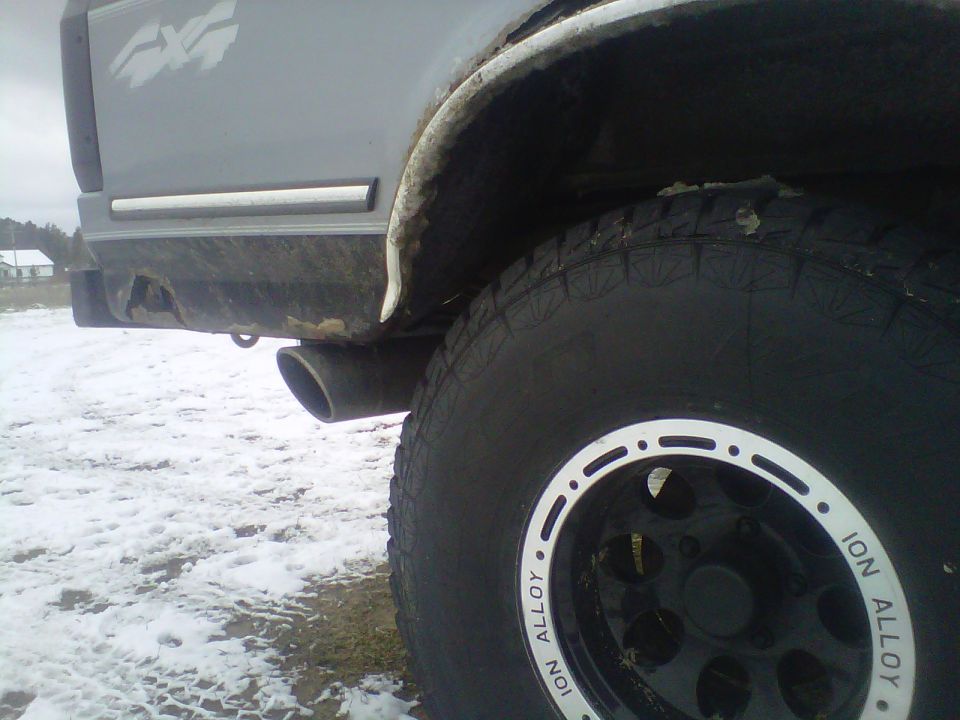 Gray truck rear quarter panel with 4x4 decal, chrome trim, and exhaust pipe. Off-road tire on black Ion Alloy wheel is prominent
