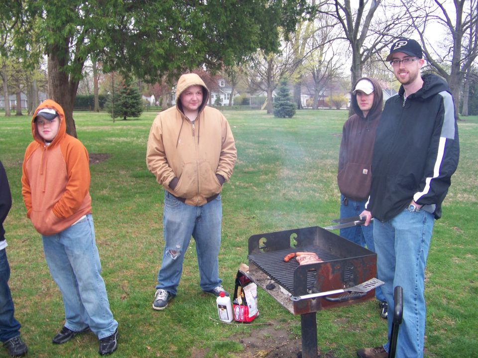 Four young men stand around a barbecue grill in a grassy park with trees in the background