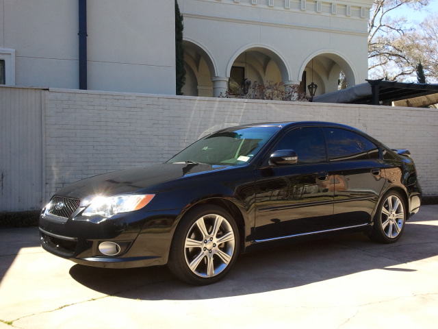 Black sedan parked in front of a white brick wall and a building with arches. The car has tinted windows and a spoiler