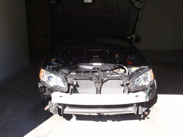 Front of a dark colored Subaru Legacy with hood open, bumper removed, showing engine and radiator