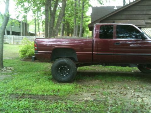 2001 Dodge 1500 - Maroon pickup truck with large tires parked in a grassy area near a wooden building.