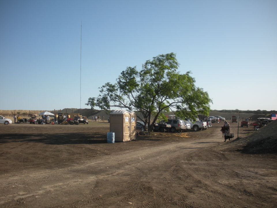 outdoor event with parked trucks, ATVs, tents, and portable toilets under a tree and tall antenna